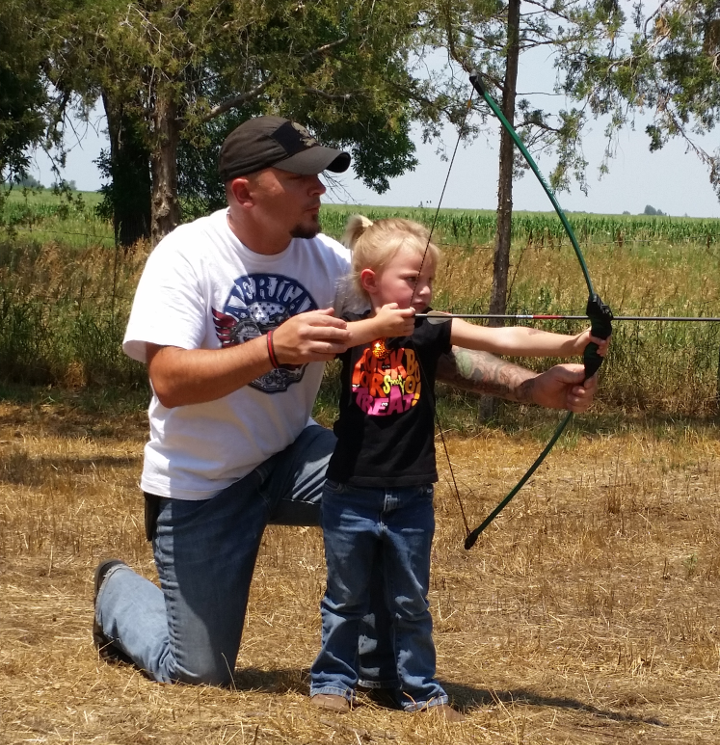 A father assists his three year old daughter in shooting an arrow by helping her pull back the bow.